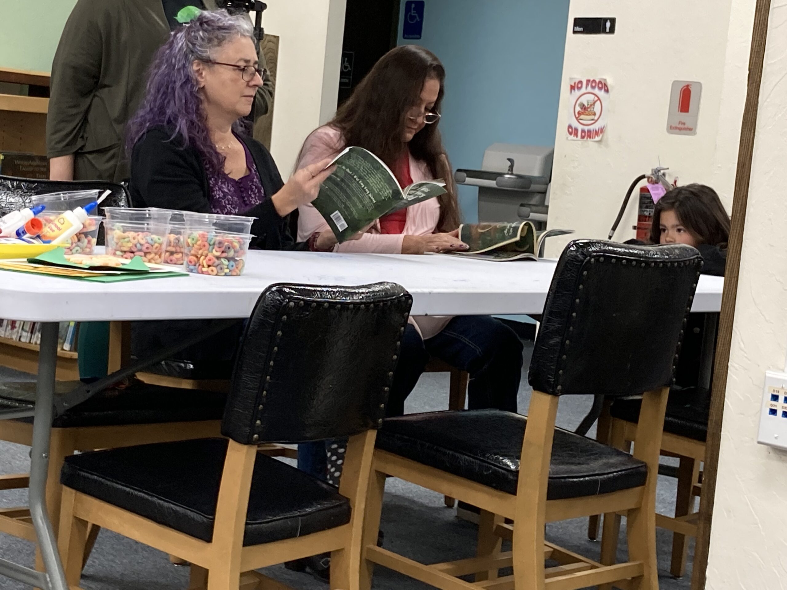 Two ladies sitting at a table reading to a young child.
